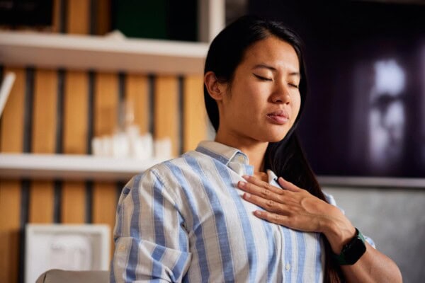 a calm asian woman sitting on a chair, holding a hand on a chest with eyes closed.