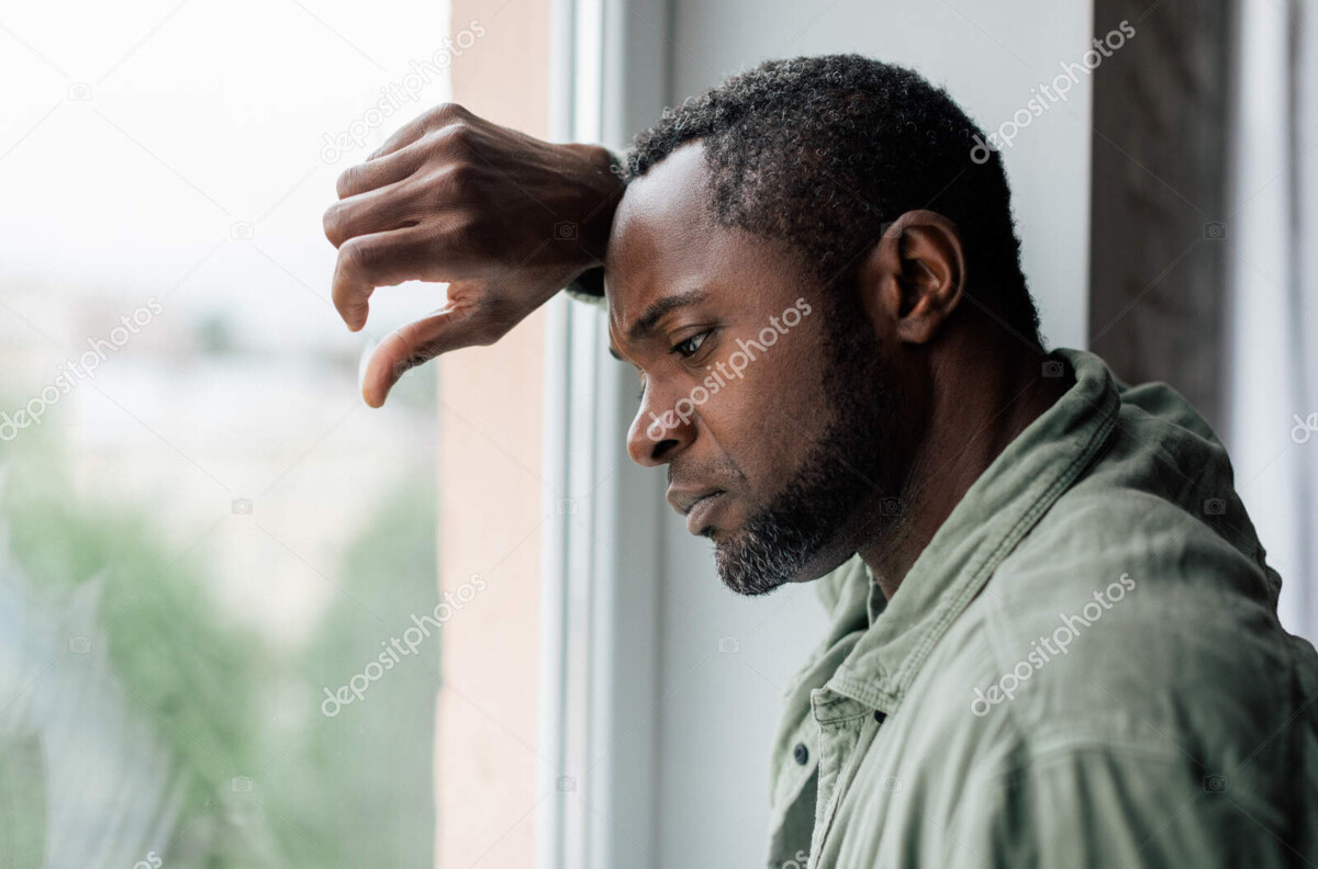 unhappy frustrated adult african american guy in casual suffering from depression and bad news near window